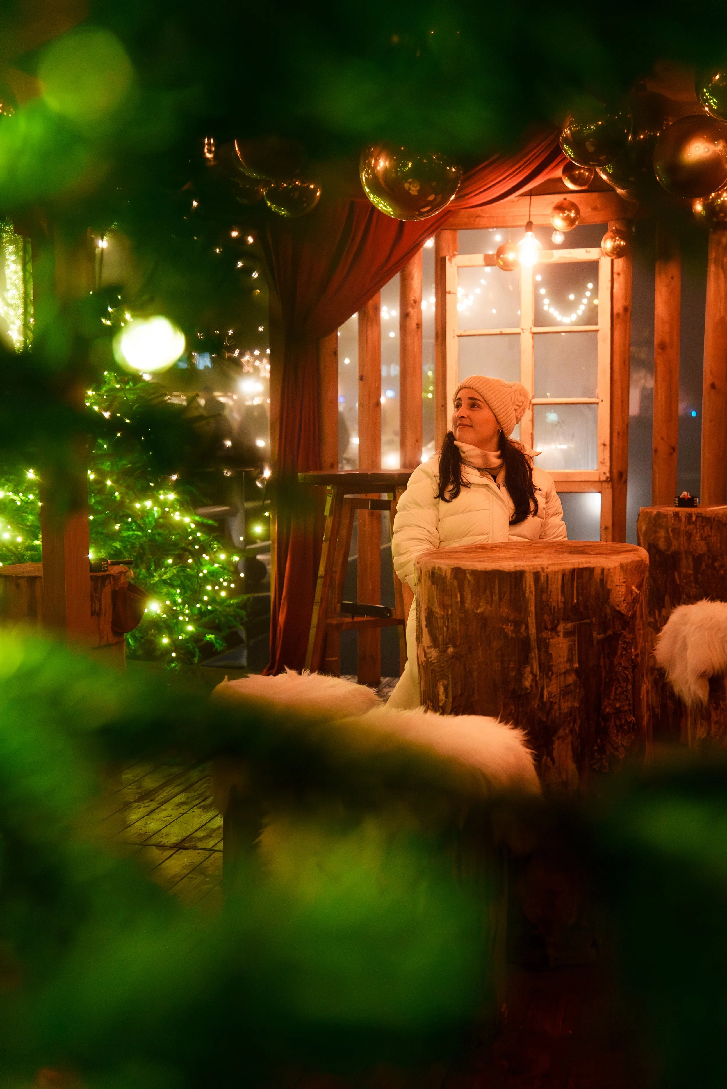 A woman in a white winter coat and pink knit hat sitting inside a cozy, decorated wooden cabin during Christmas, with string lights and Christmas trees visible.