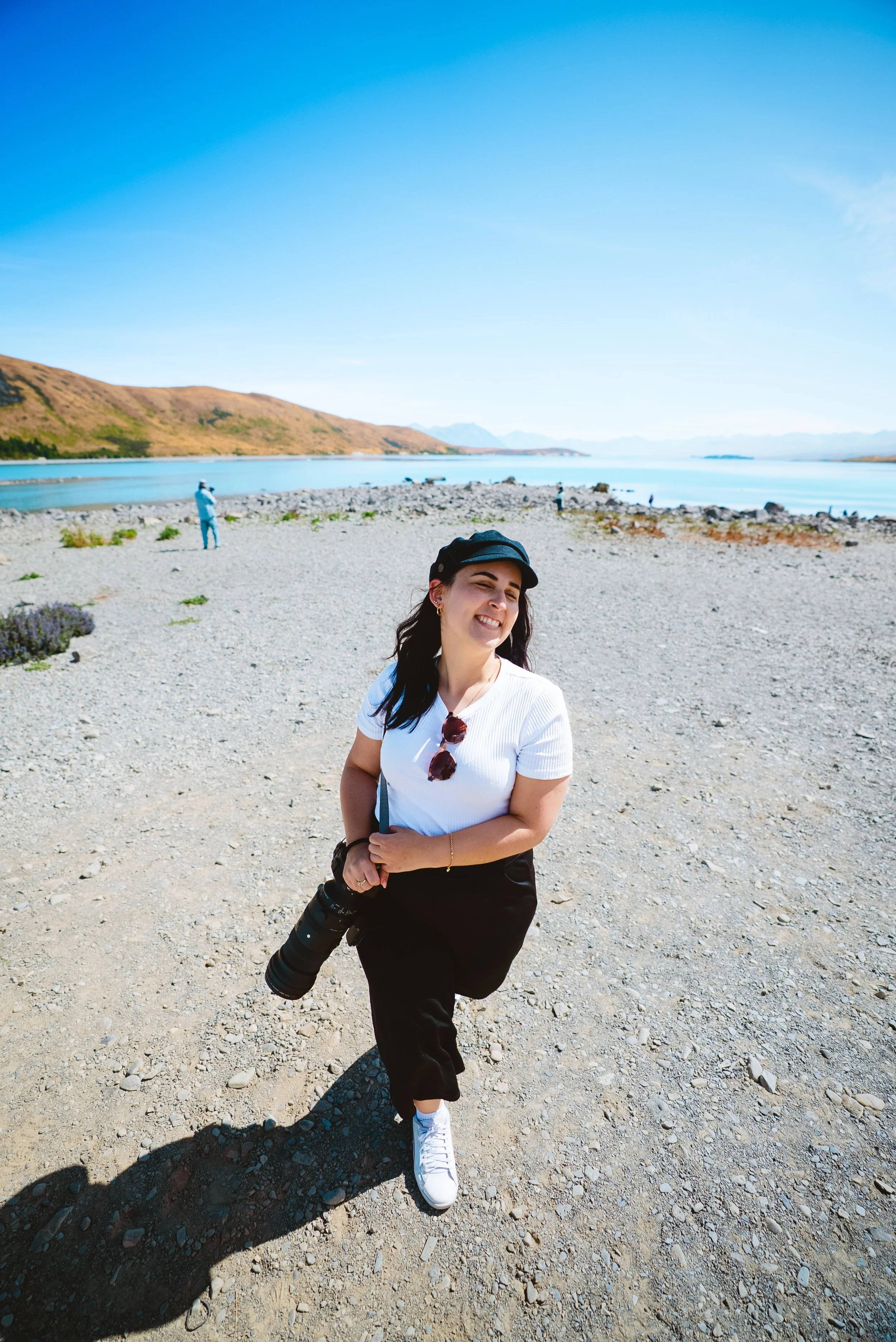 Young woman standing on a rocky beach, smiling, holding a camera, with a scenic lake and mountains in the background.
