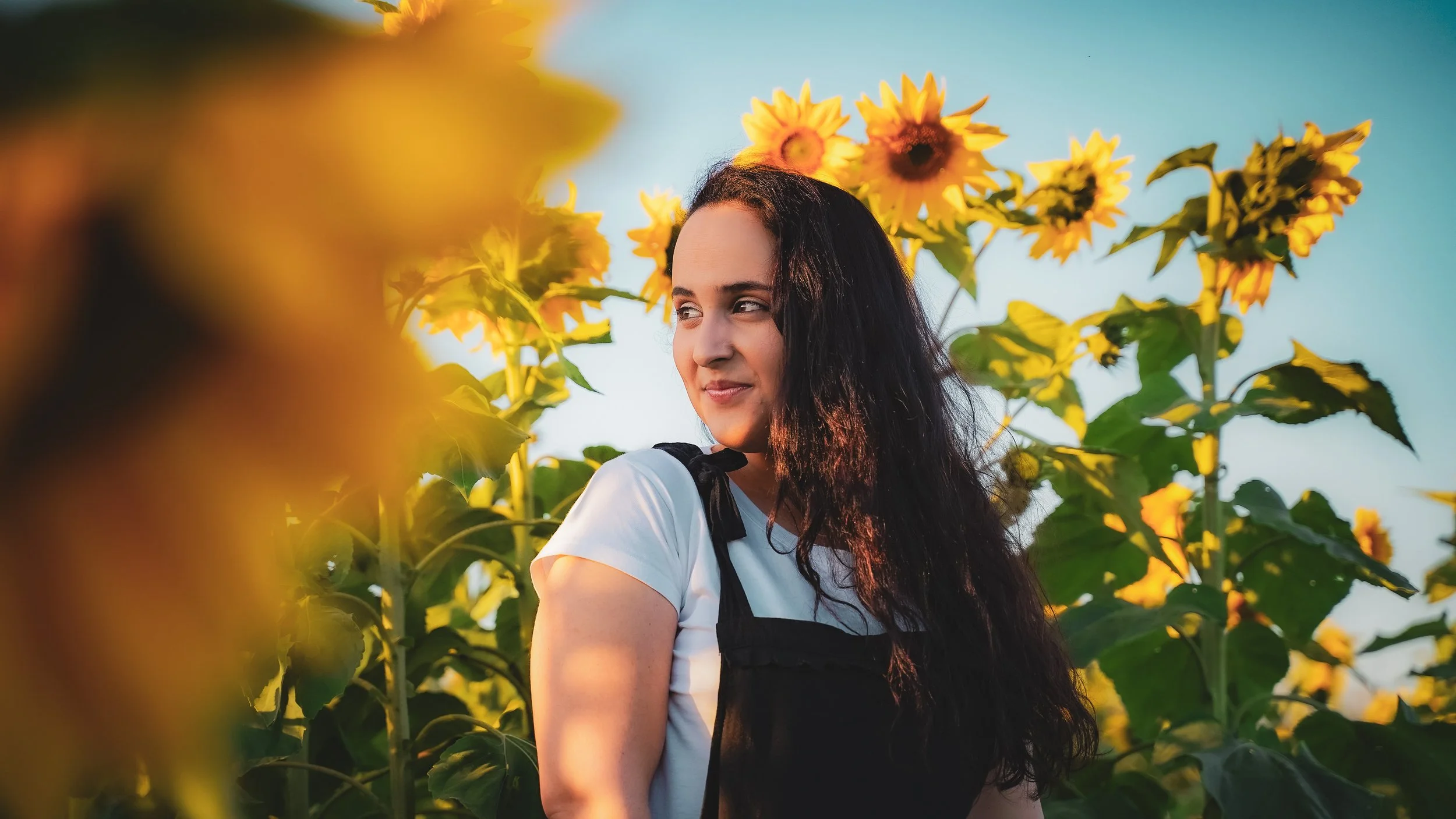 A young woman with long dark hair standing among tall sunflowers on a sunny day, looking to her right with a gentle smile.