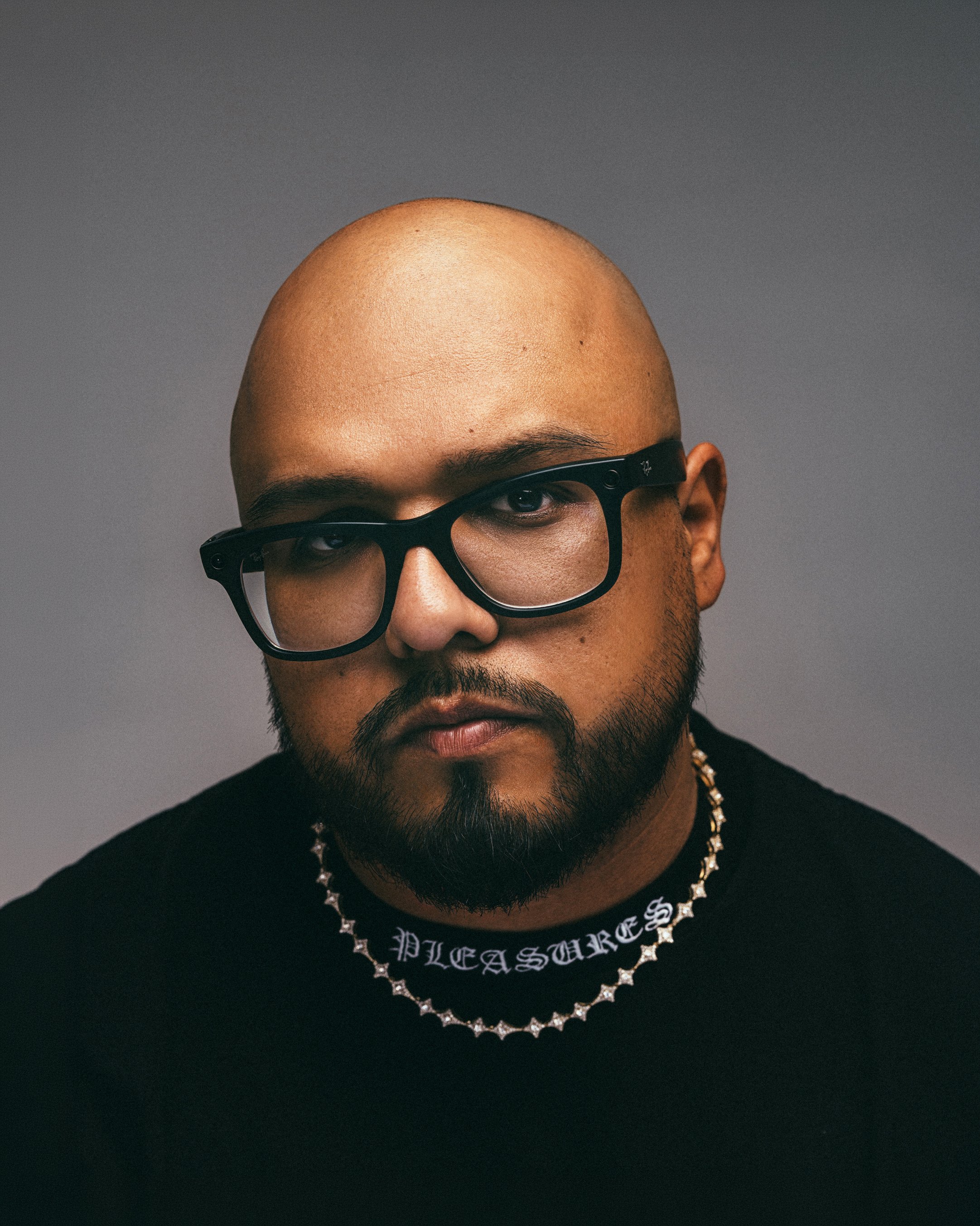 A man with a shaved head wearing thick black glasses, a black shirt, a silver chain necklace, and a black necklace with the word 'PLEASURE' on it, against a gray background.