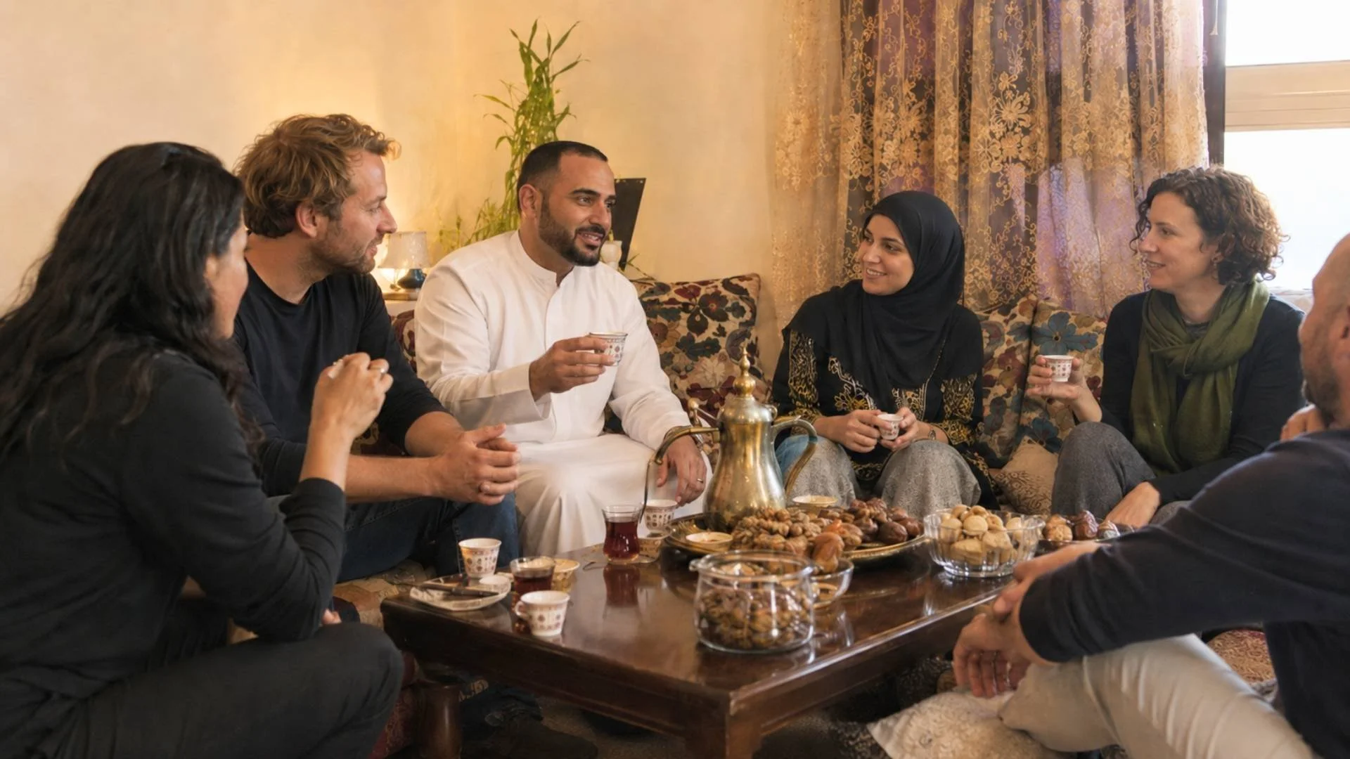 Arab hosts and non-Arab guests sharing Arabic coffee and sweets in a warm home setting.