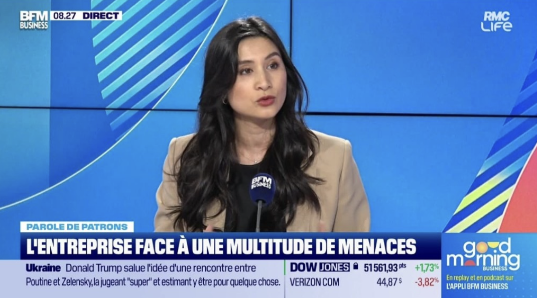 Femme en interview lors d'une émission de télévision avec une veste beige, tenant un micro évoquant BFM Business, à l'écran des graphiques et textes liés à l'économie et aux menaces contre les entreprises.