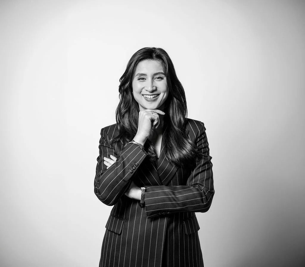 Une femme souriante en costume à rayures noires et blanches, les bras croisés, posant contre un fond clair.