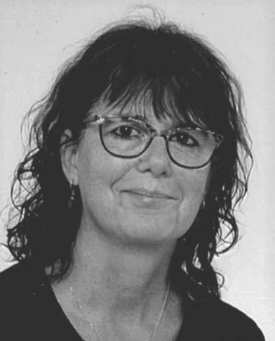 Schwarz-weiß-Foto einer Frau mit dunklen, lockigen Haaren, trägt Brille und einen schwarzen Pullover.