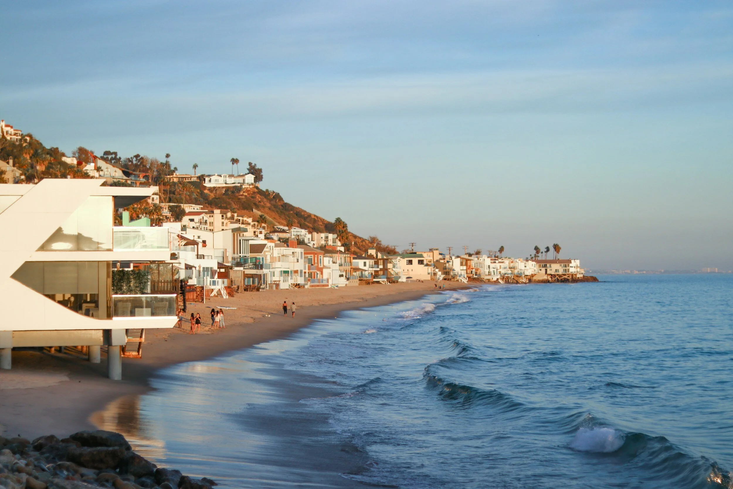 A scenic view of a beach with modern houses on a hillside, calm ocean waves, and a few people walking on the sandy shore during sunset.