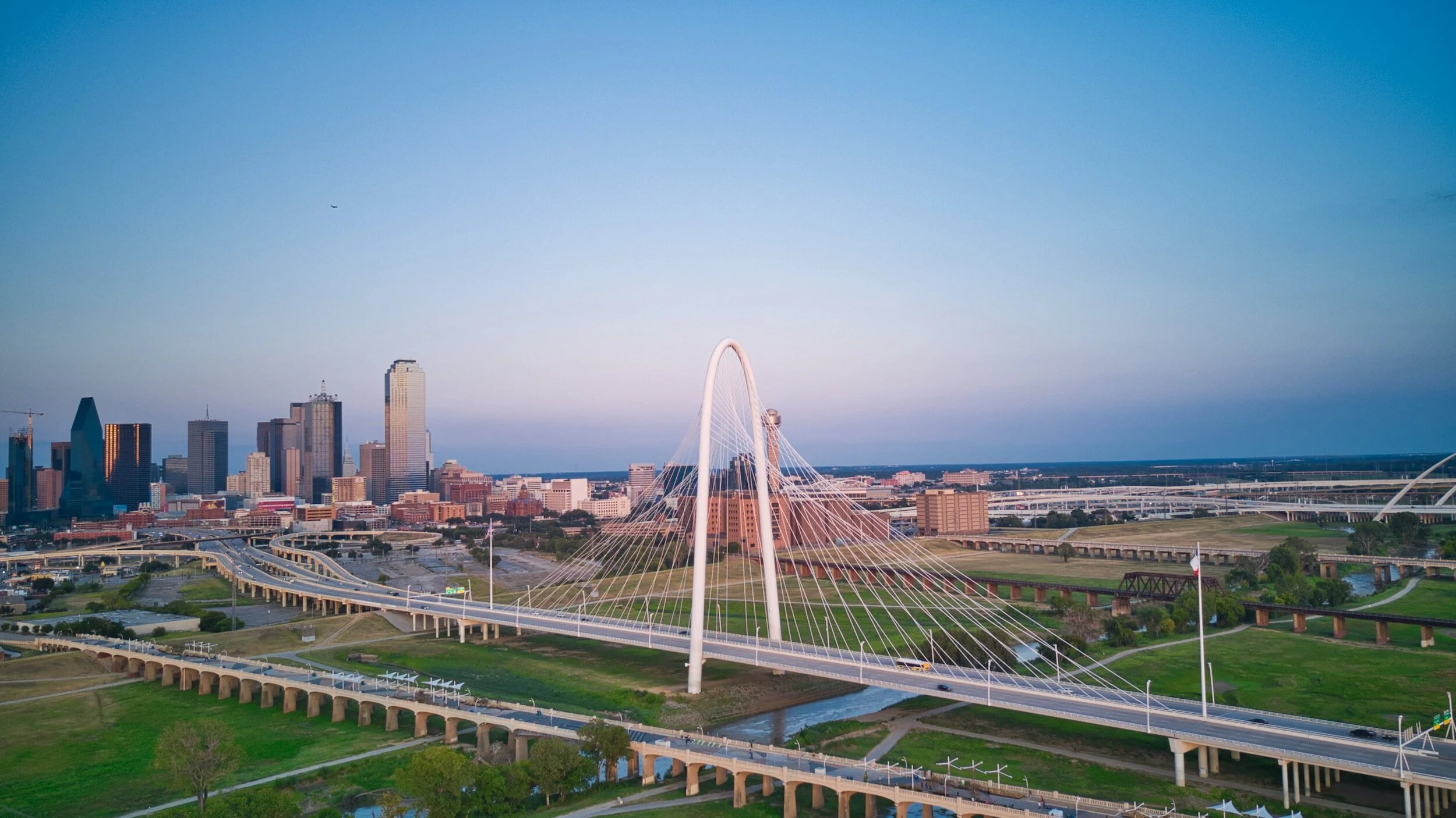 Dallas skyline with Margaret Hunt Hill Bridge in foreground, green parks, and highways under a clear sky