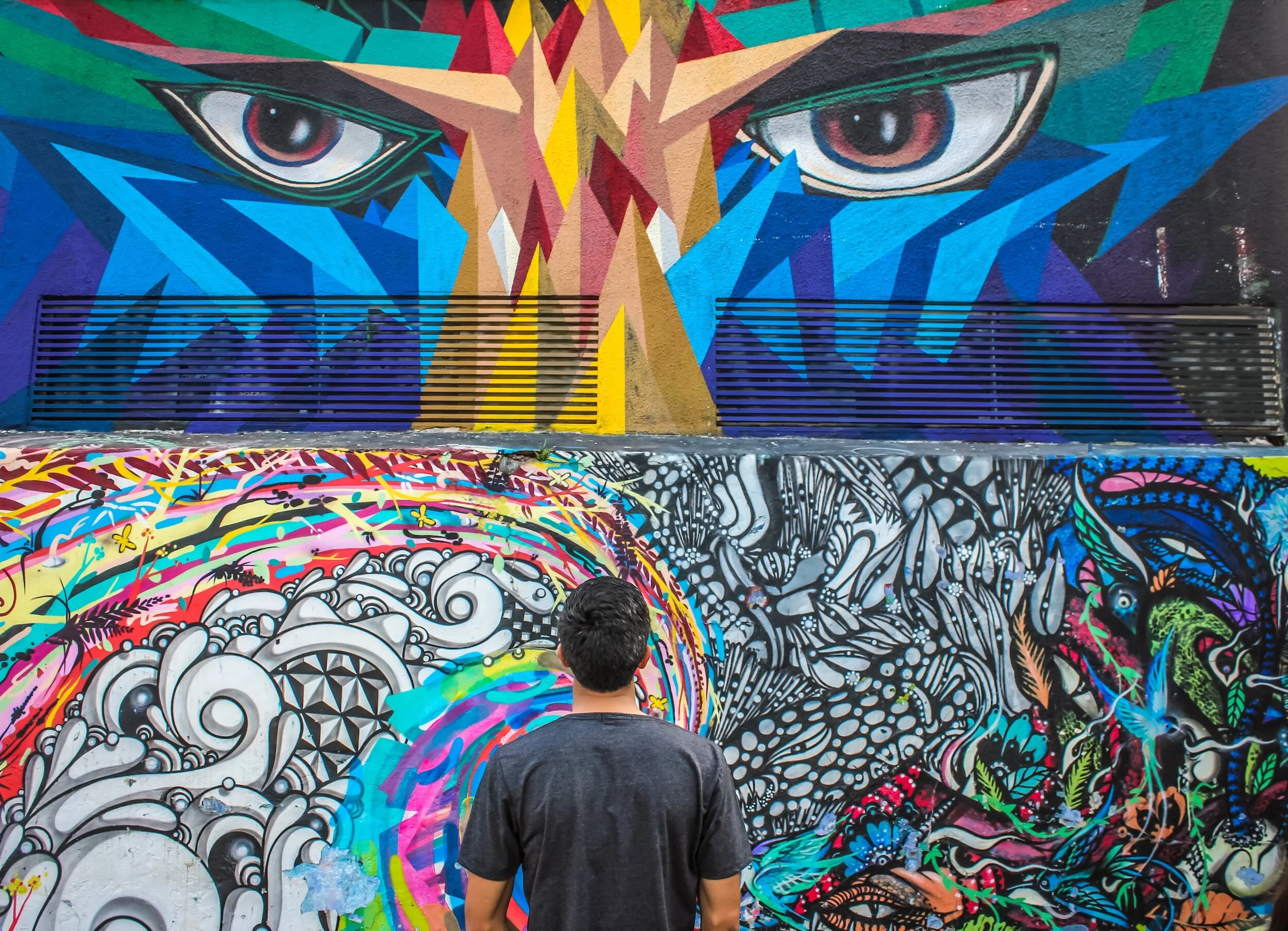 A man standing in front of colorful graffiti art on a wall, featuring a large, fierce face with intense eyes at the top and intricate, vibrant abstract patterns below.