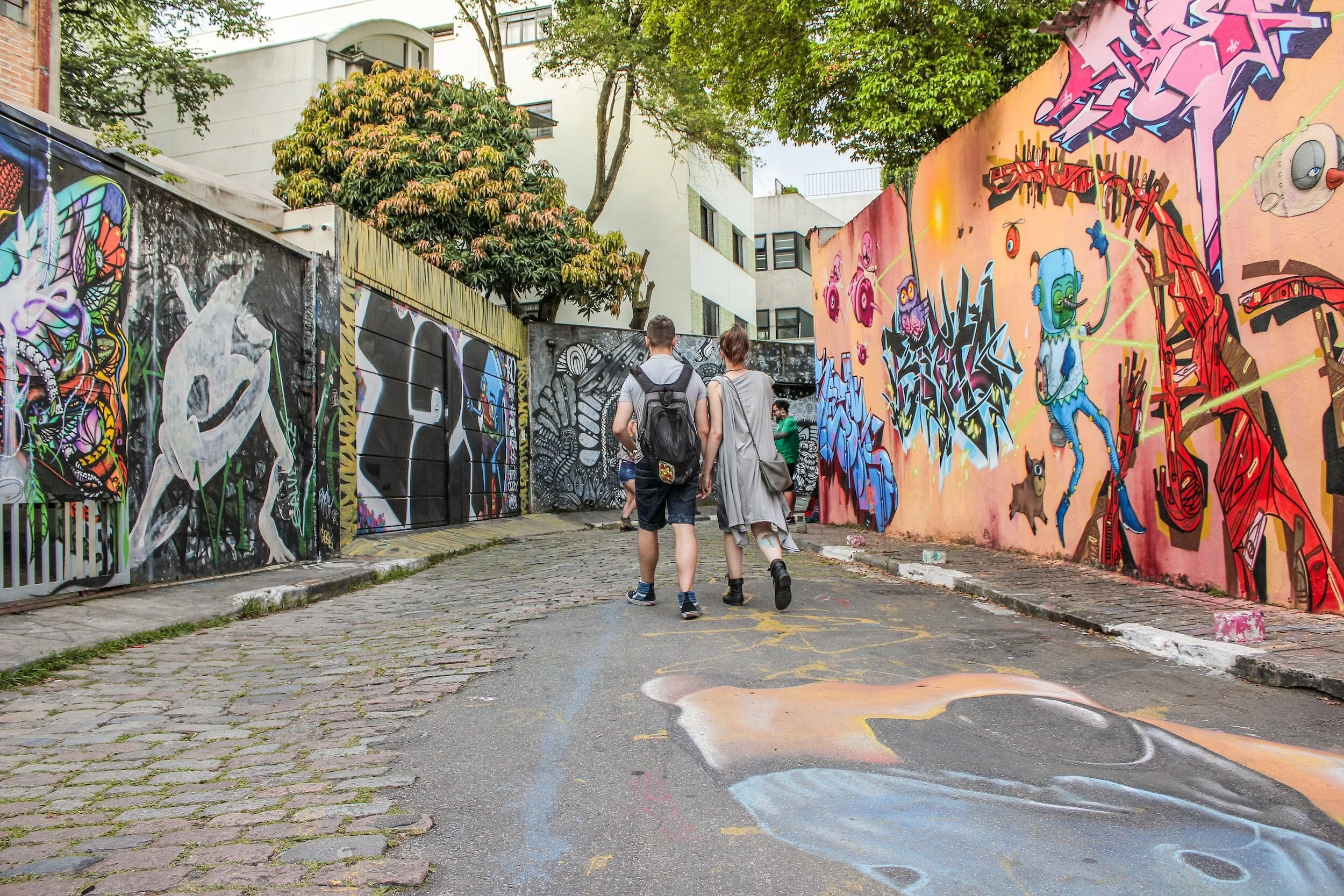 Two people walk down a narrow, cobblestone alleyway with colorful graffiti murals on both sides, featuring various vibrant abstract and character designs.