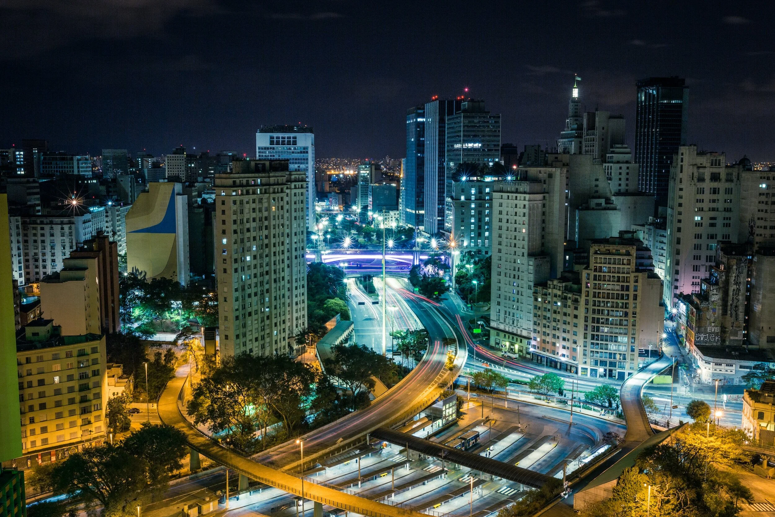 Night view of a city skyline with illuminated skyscrapers, roads with light trails, and trees.