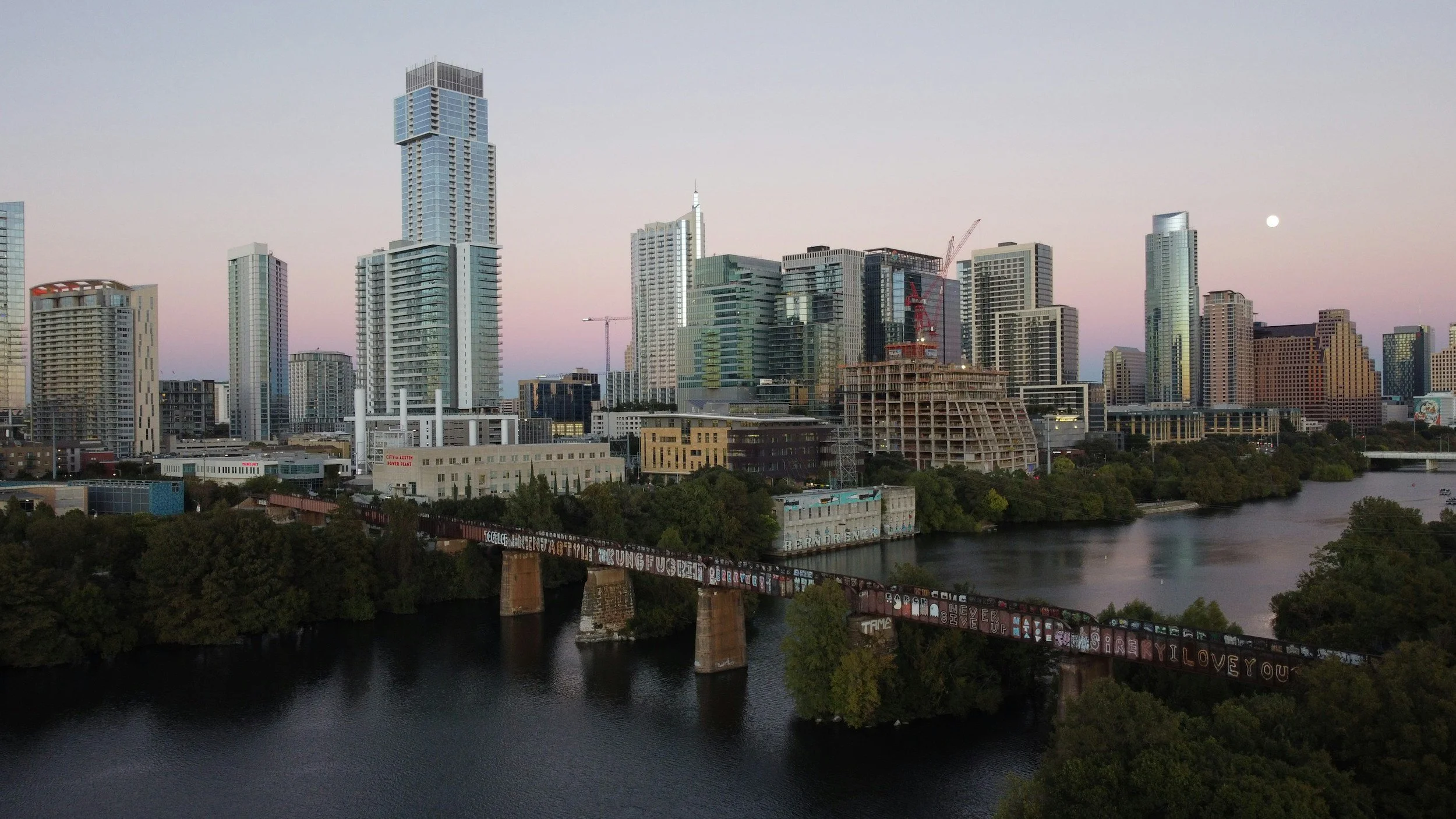 City skyline of downtown Austin, Texas with tall modern buildings, a river in the foreground, and graffiti-covered train tracks crossing the river. The sky is light with a visible moon.