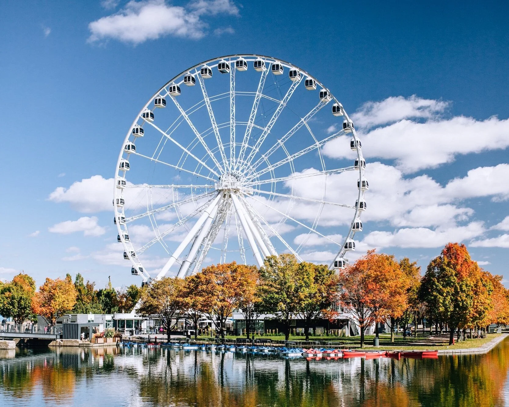 A large white Ferris wheel near a body of water, with trees displaying fall colors and a blue sky with scattered clouds in the background.