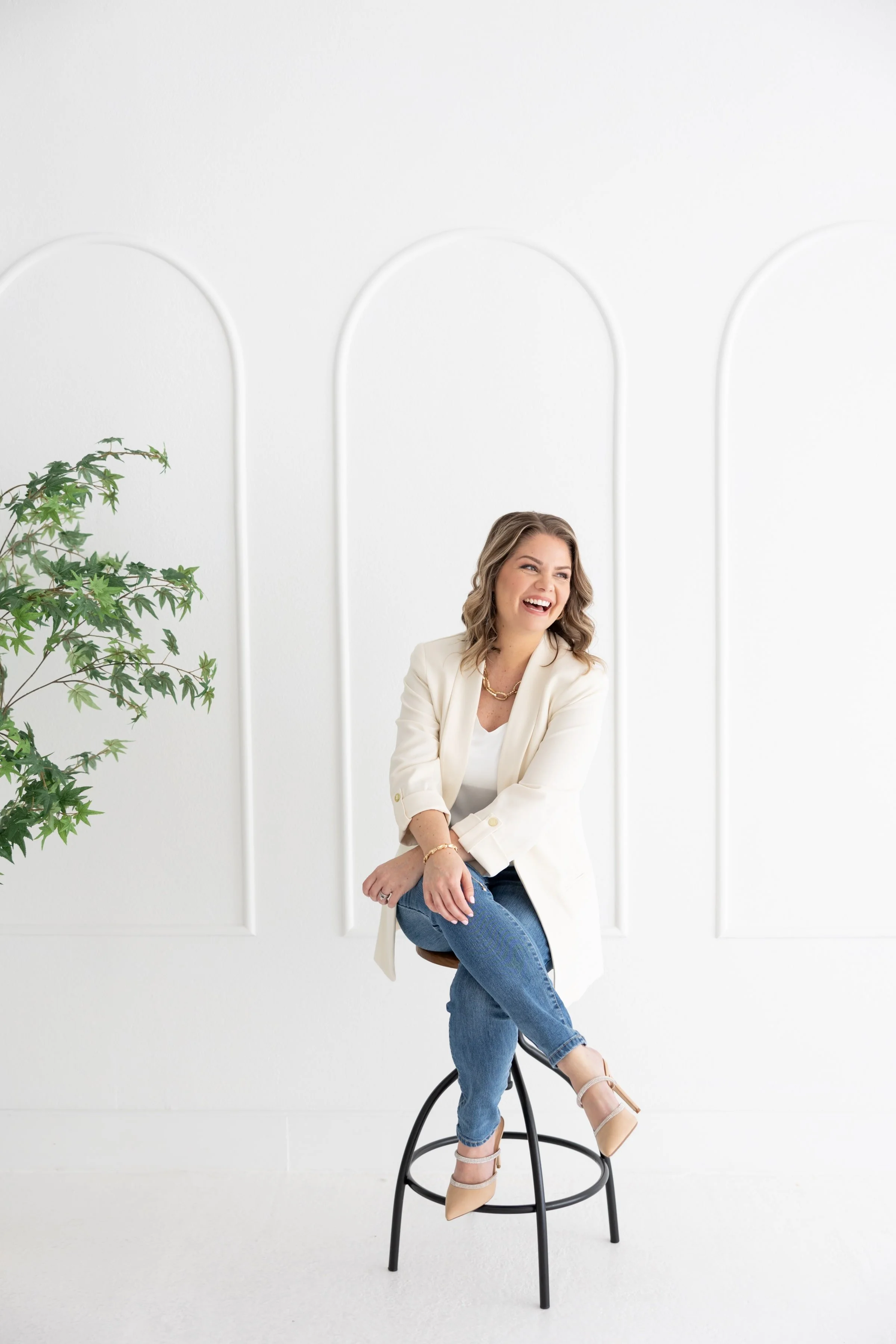Woman sitting on a stool in a white room, smiling and wearing a white blazer, jeans, and beige heels.
