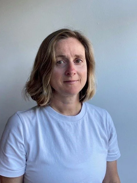 A woman with shoulder-length wavy hair wearing a white T-shirt standing against a light-colored wall.