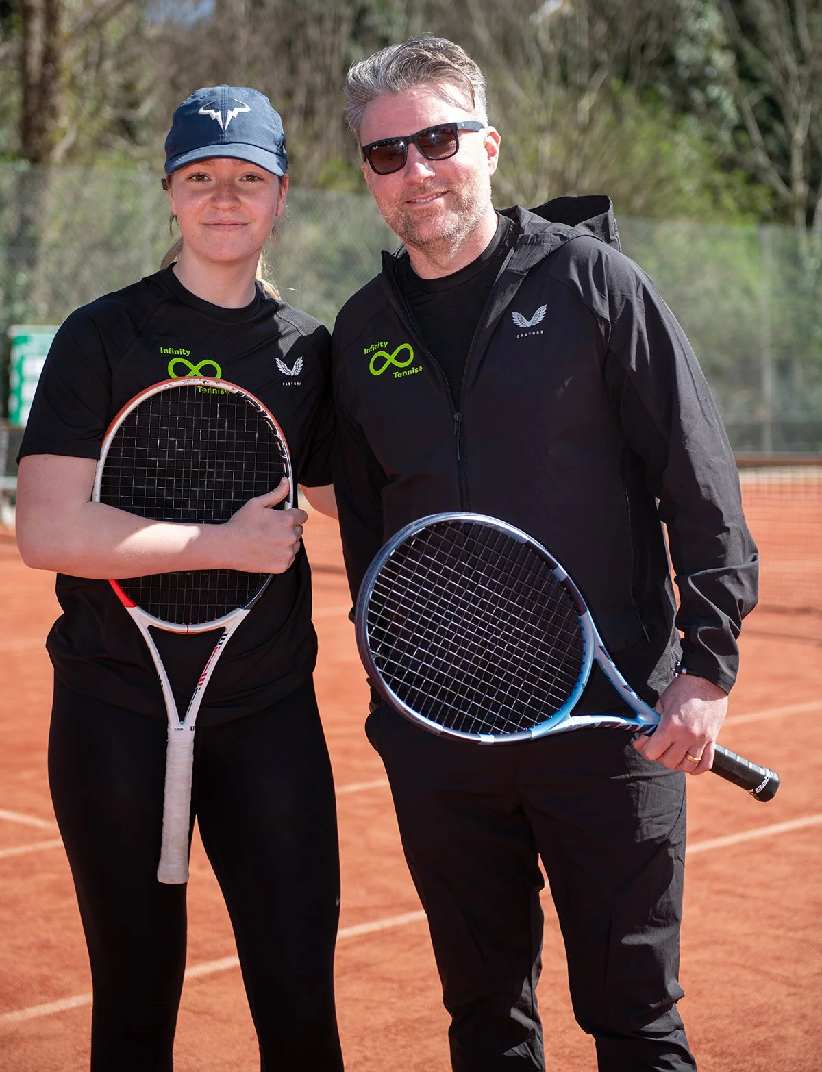 Mia and Gavid standing side by side on a tennis court, both holding tennis rackets, smiling at the camera, wearing black sports clothing with a logo, and the young woman is wearing a blue cap.