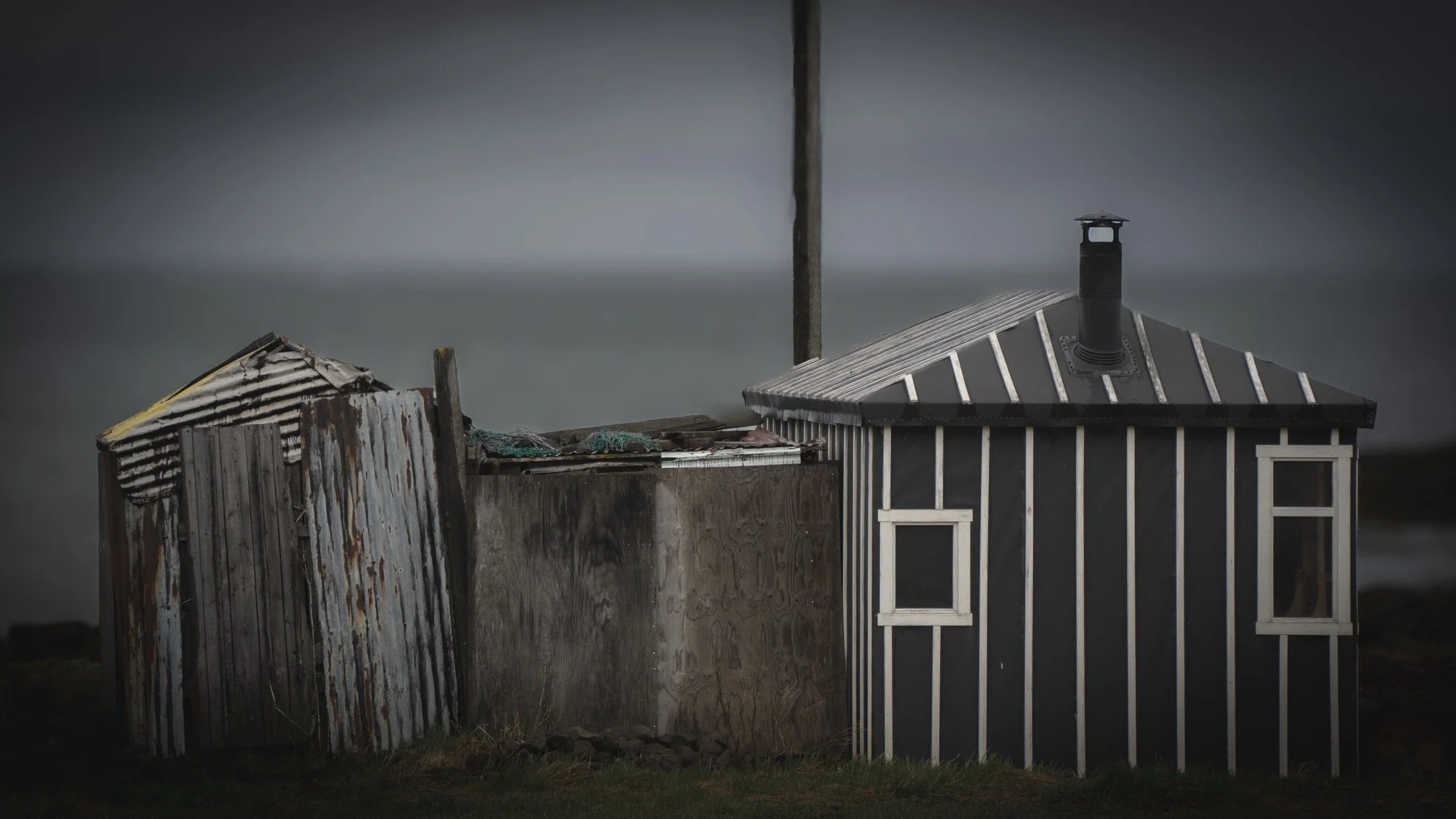 Two small houses with contrasting appearances; one old and weathered with rusty, peeling metal siding and a damaged roof, and the other modern with fresh black and white striped walls and a functioning chimney, set against a dark, cloudy sky.
