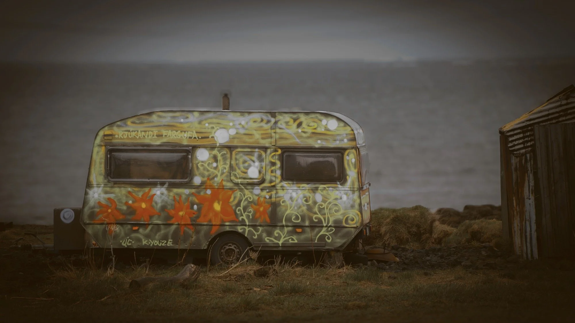 A colorful vintage camper trailer with orange and yellow floral designs parked on a grassy area near water, with a small wooden shed nearby and cloudy sky overhead.