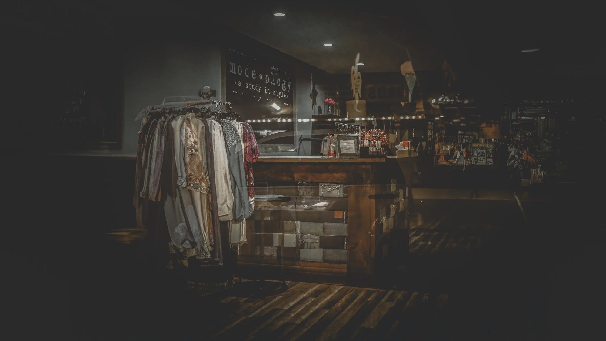 A clothing store with a rack of hanging garments in the foreground, a cash register desk behind it, and shelves filled with various items in the background, all under dim lighting.