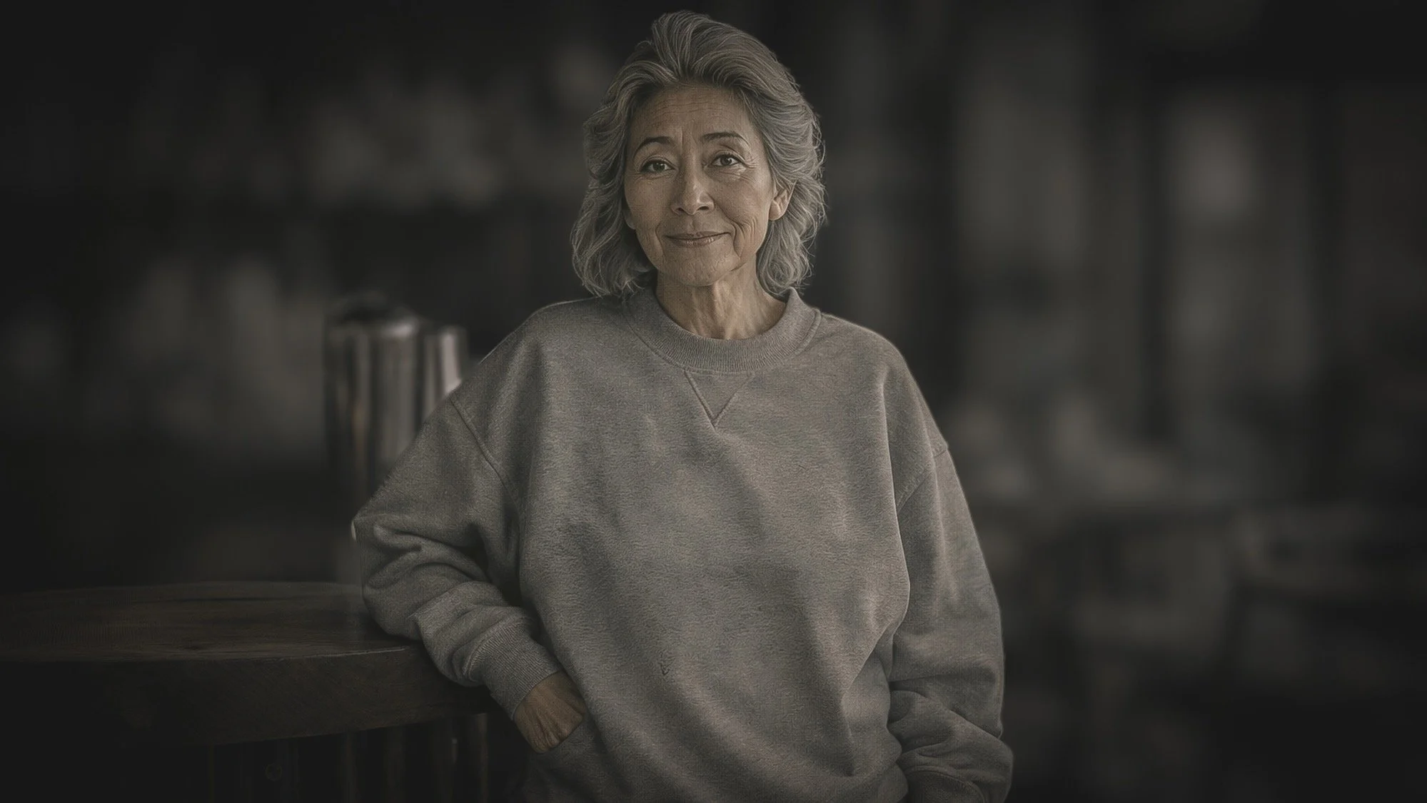 An older woman with gray hair, wearing a gray sweatshirt, standing indoors in front of a blurred background, smiling softly.
