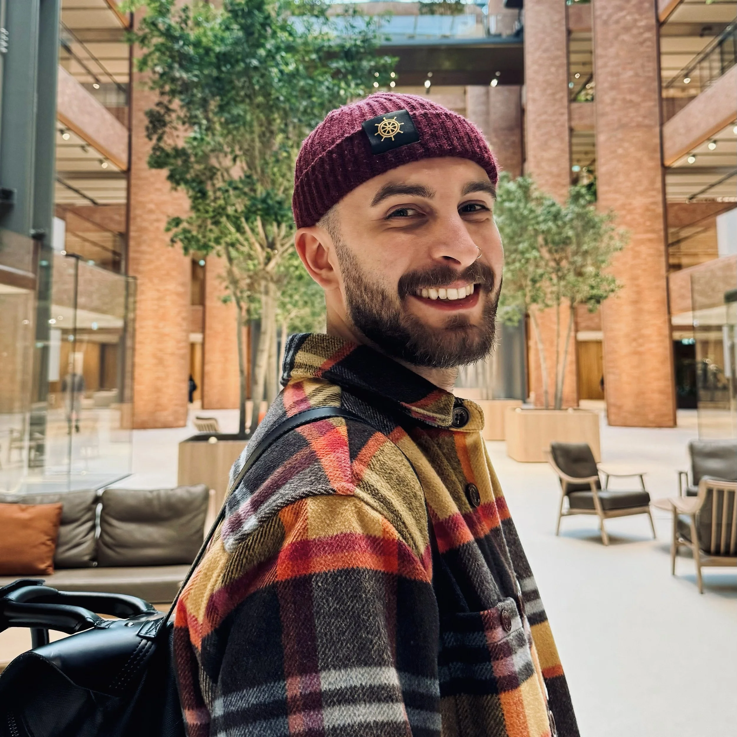 A man wearing a burgundy knit beanie with a yellow nautical emblem, smiling, in a modern indoor setting with brick walls, glass railings, and trees.