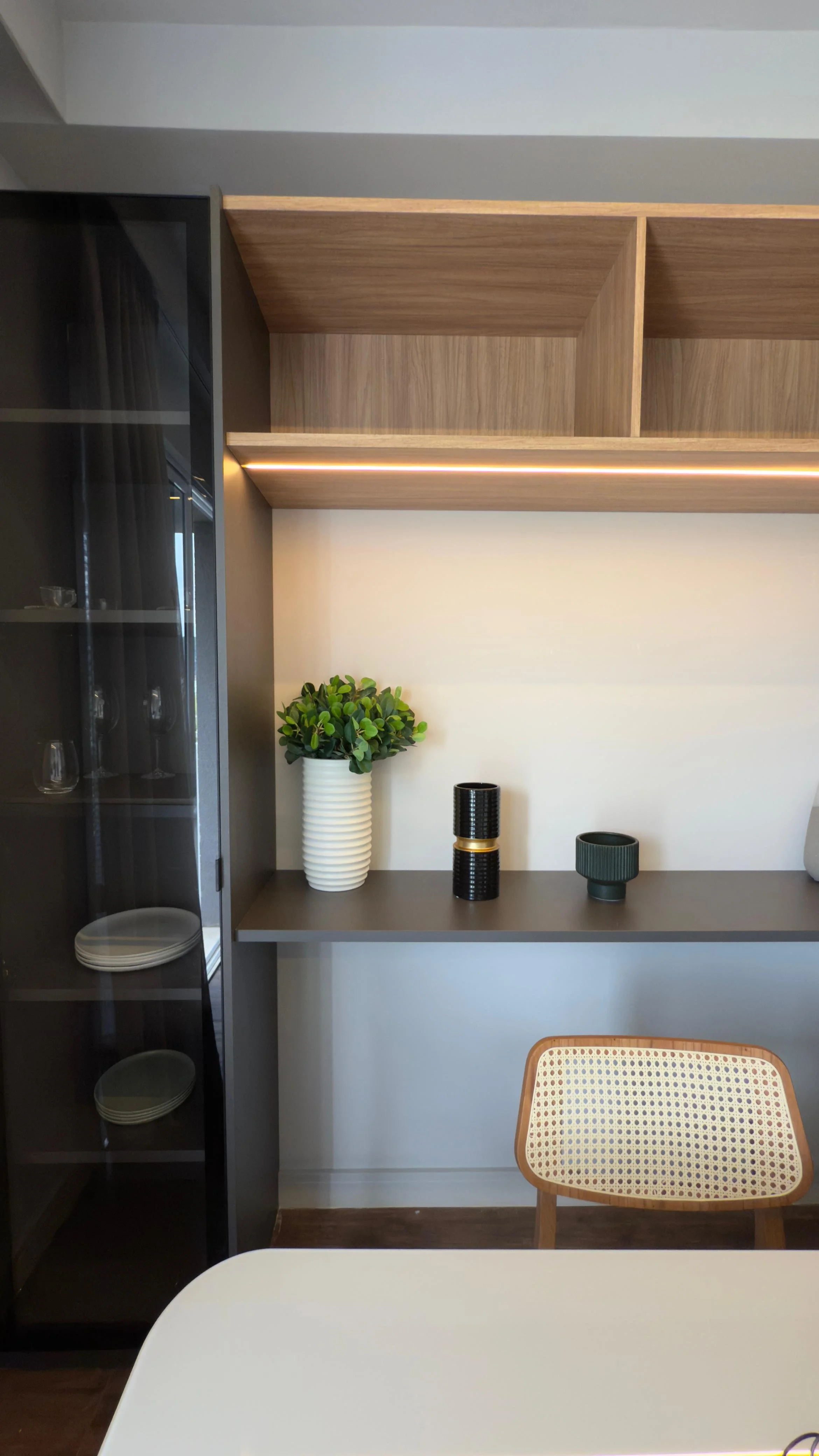 Sala de jantar com mesa redonda branca, quatro cadeiras cinza, prateleira com nichos e geladeira preta, cortinas de tecido transparente na janela, piso de madeira.