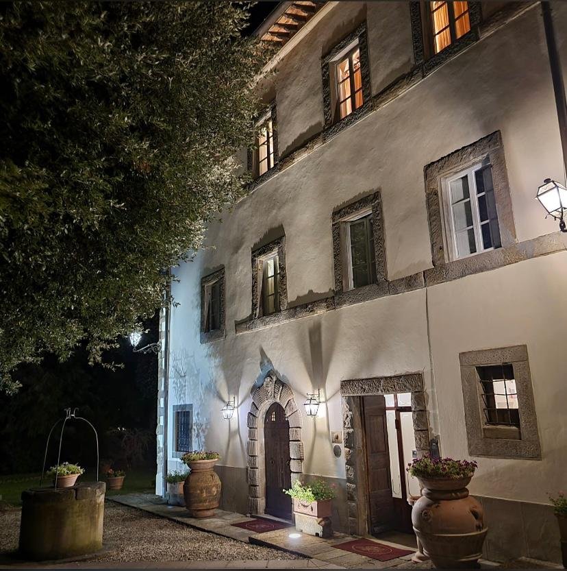 Et gammelt, fire-etasjers hus med lysende vinduer og utendørs belysning om kvelden. Det er store potteplanter ved inngangen og en brostein-dør med lysene ved siden av.