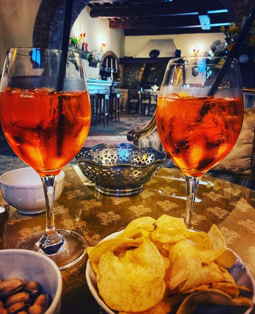 To glass med rosa drikke, en skål med potetgull, og en annen liten skål med nøtter på et bord med dekke, i et rustikt interiør.