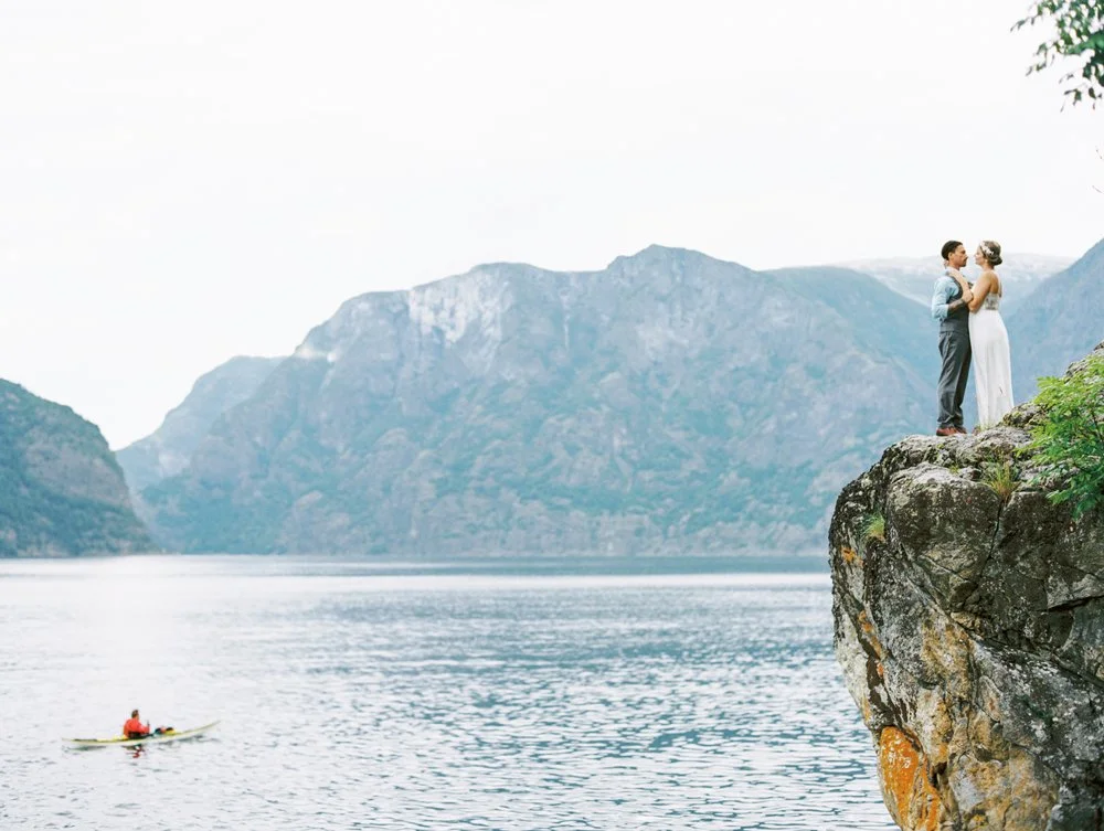 Et brudepar i brudekjole og dress står på en klippe ut mot sjøen med fjell i bakgrunnen, mens en person padler forbi i kano.