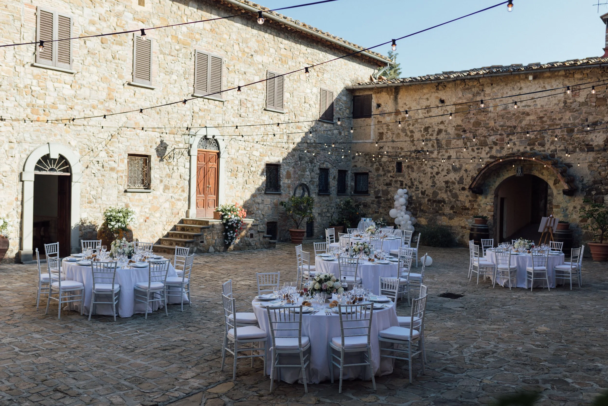 Utendørs bryllupsfeiring med flere hvitkledde bord, pyntet med blomster, på en rustikk gårdsplass i Umbria. Området er pyntet med vakre lyslenker og blomsterdekorasjoner.
