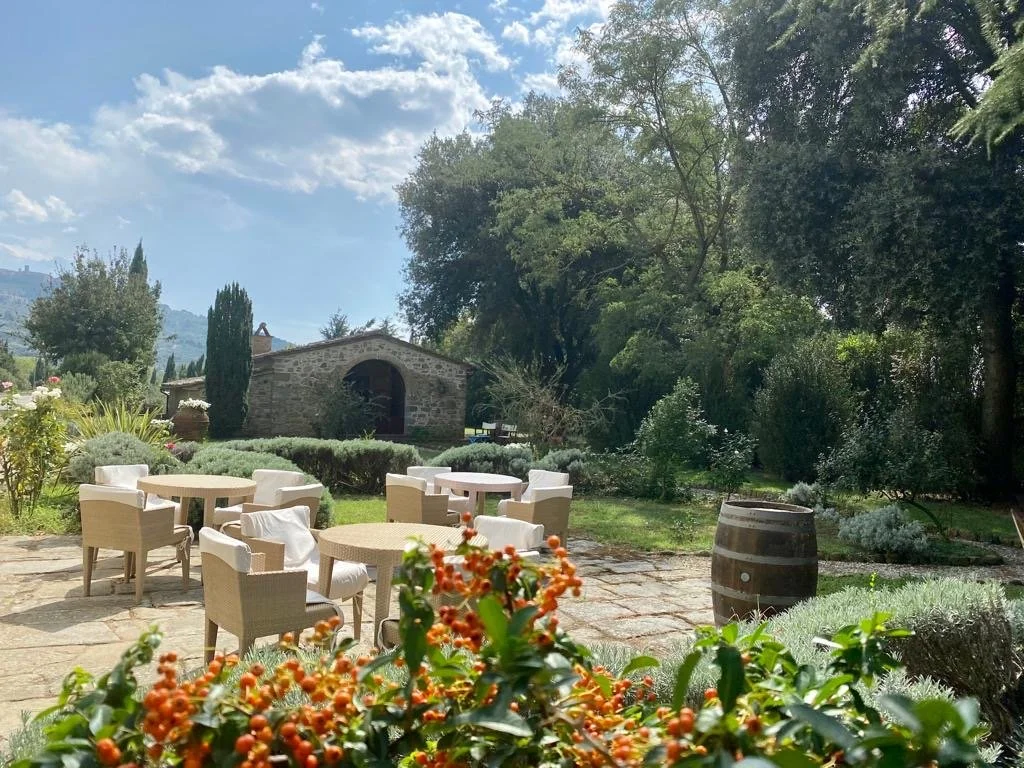 Utendørs hage med stoler og bord, omgitt av grønne busker og trær, med en steinbygning i bakgrunnen. Solfylt dag.