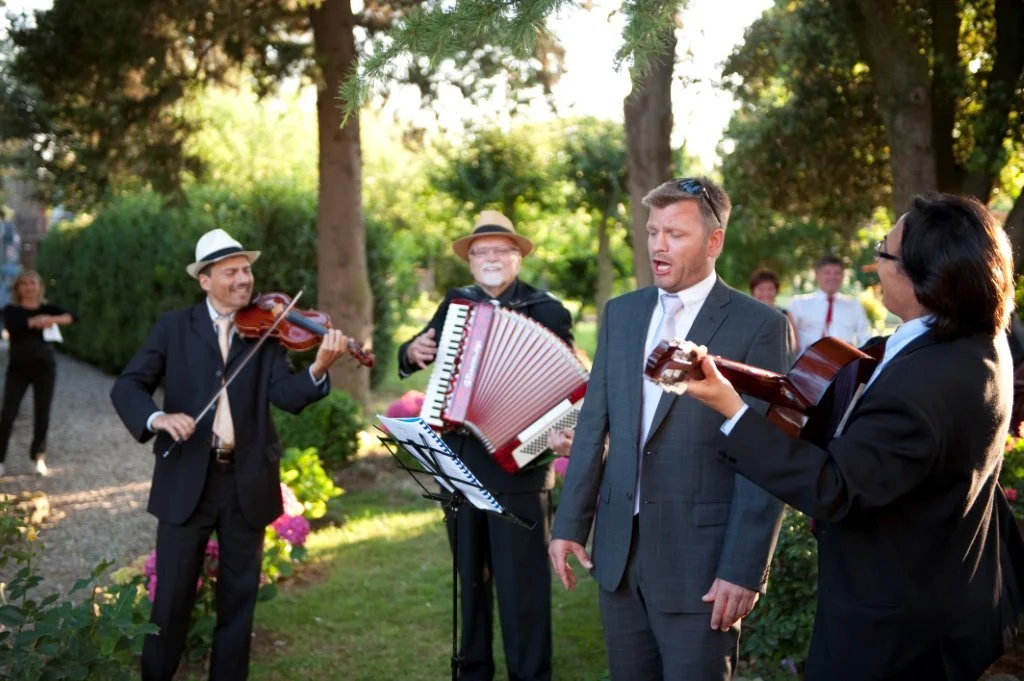 Mann synger mens to andre spiller fiolin, gitar og trekkspill i en hage.