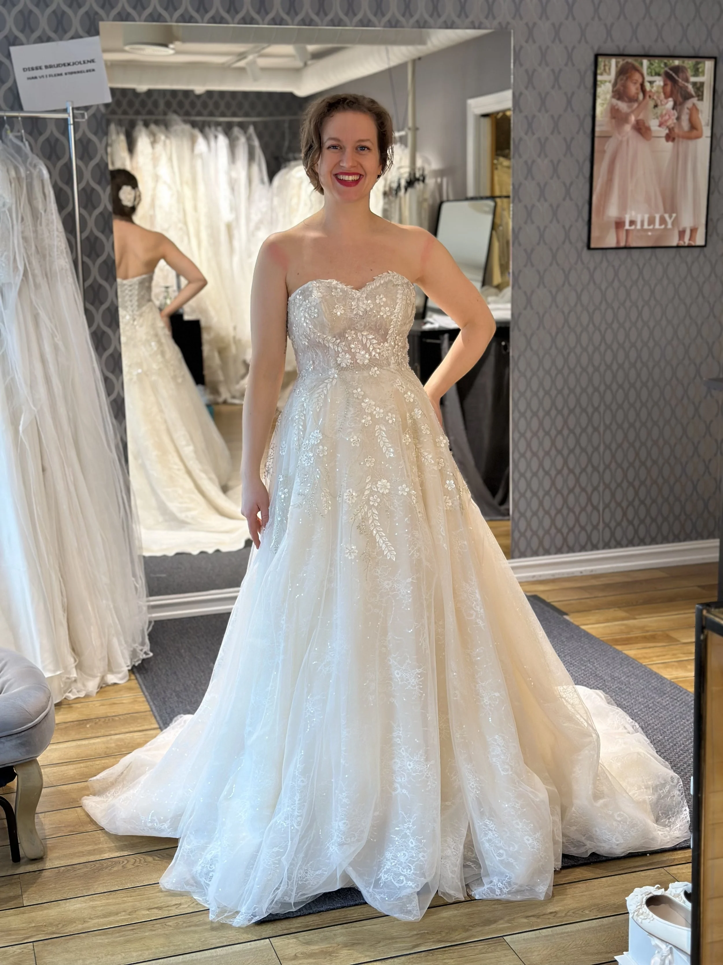 Nydelig tylldrøm med tubetopp og snøring i ryggen. Brudekjolen er i fargen nude, og har glitrende sølvfargede detaljer med paljetter og påsydde blomsterapplikasjoner i ivory. Denne brudekjolen er fra Lilly og er en av våre nye brudekjoler