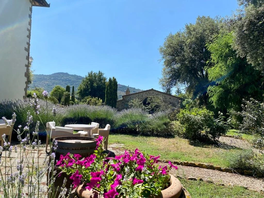 En hage med blomster, sitteområder med hvite stoler og bord, og en liten bygning med steinmur i bakgrunnen, omgitt av grønne trær og busker under en klar blå himmel.