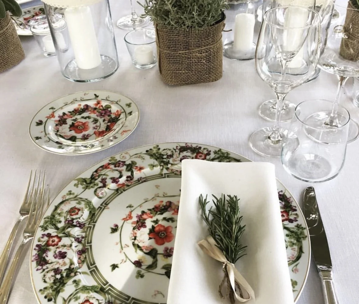 Festbord med floralmønstrede tallerkener, bestikk, glass, en hvit serviett med en små kvist rosmarin, og dekorative lyslykter og små grønne planter i bakgrunnen.