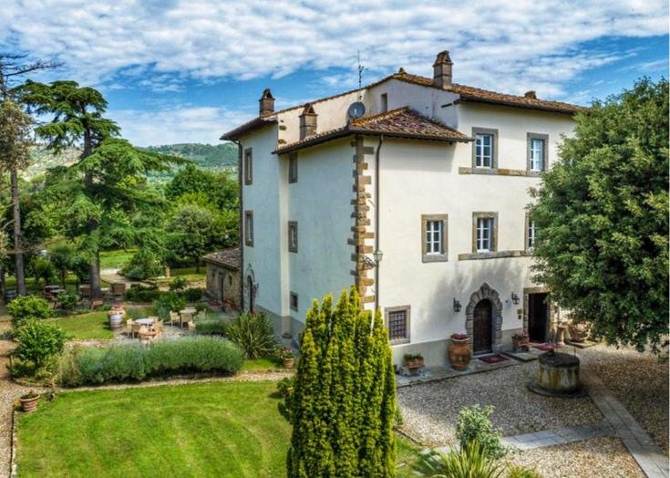 En hvit, tre etasjers villa med tåkonstruksjon, omgitt av frodige grønne planter, trær og en velstelt hage med sitteplasser. Bildet er tatt på en klar dag med blå himmel og noen skyer.