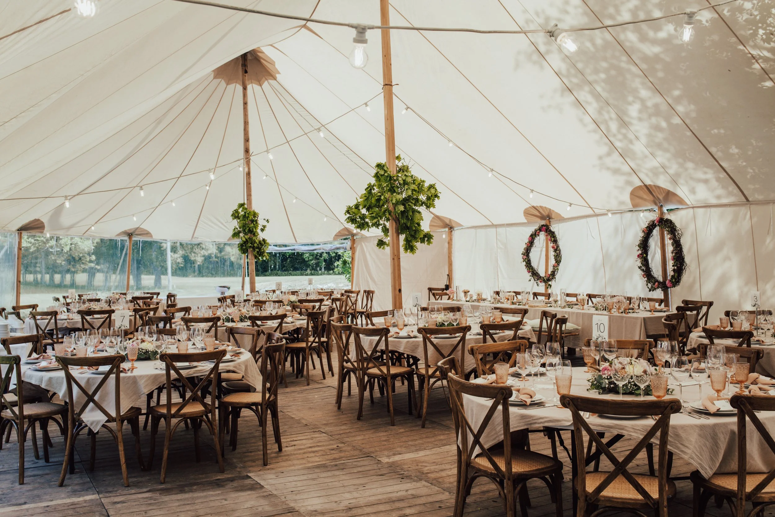 Bryllupstelt pyntet med vakre dekkede langbord og runde bord, blomsterarrangementer, glass og servise. Dekorative blomsterkranser henger på veggen. Lyslenker henger fra taket.