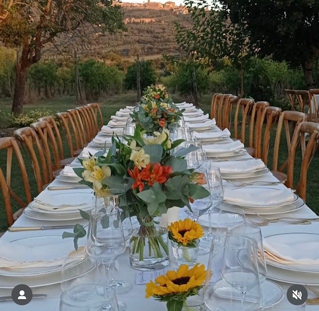 Langbord dekket med hvite duker, blomsterbuketter, glass, tallerkener og bestikk, satt opp utendørs i en hage med trær og åser i bakgrunnen.