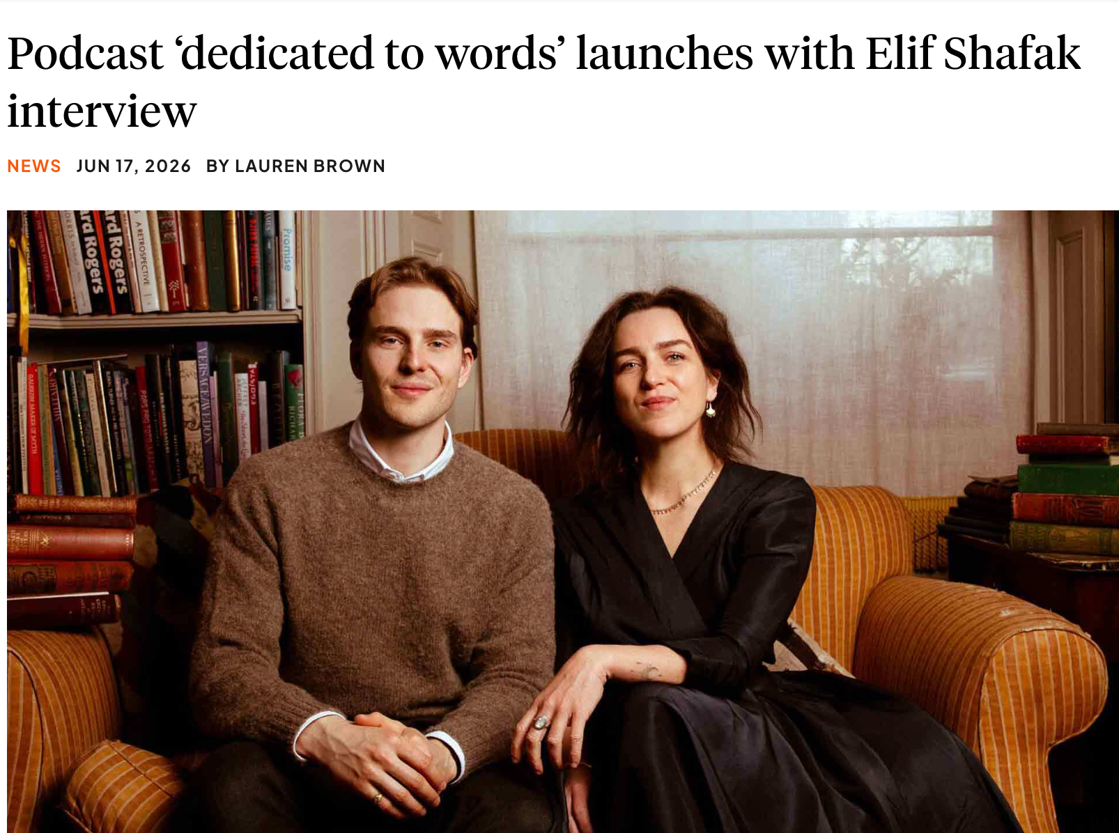 Two people sitting on a yellow striped sofa in a cozy room with a bookshelf filled with books behind them. The man on the left has brown hair, wearing a brown sweater with white shirt collar visible underneath. The woman on the right has dark curly hair, wearing a black dress and jewelry. They are looking at the camera.