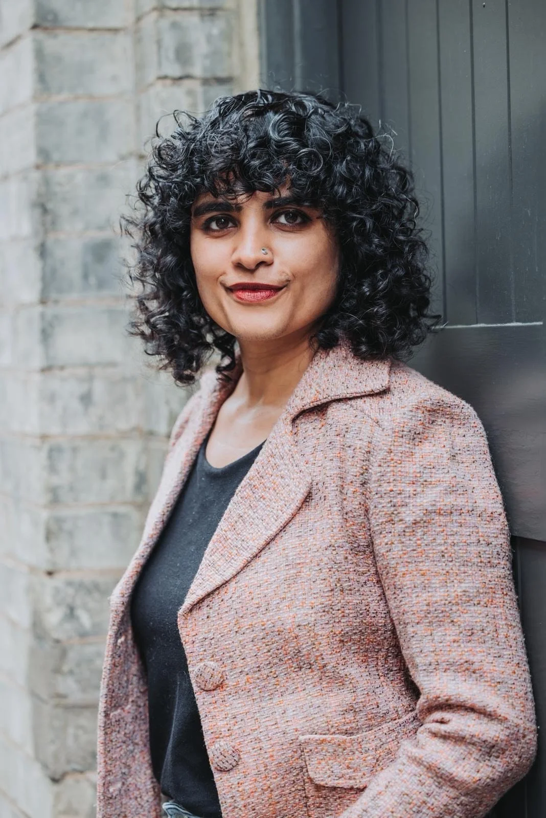 A woman with curly black hair and a nose piercing standing outdoors against a brick wall and dark wooden door, wearing a peach-colored coat over a black top.