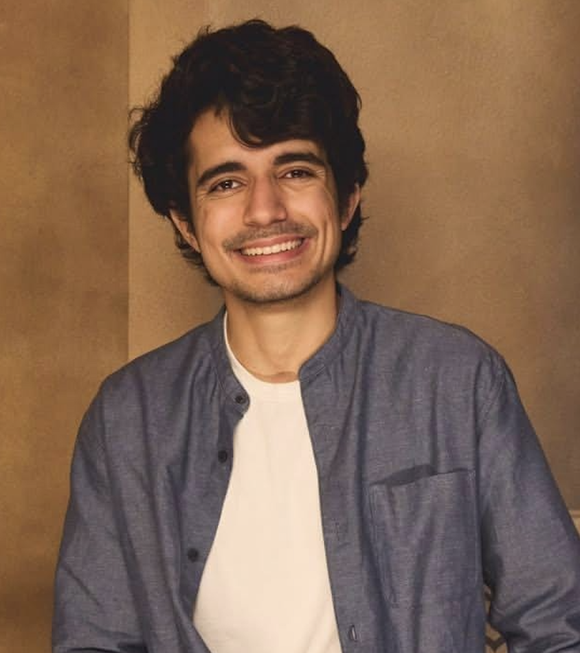 A young man with dark curly hair, smiling and wearing a white t-shirt under a buttoned gray shirt, standing against a brown background.