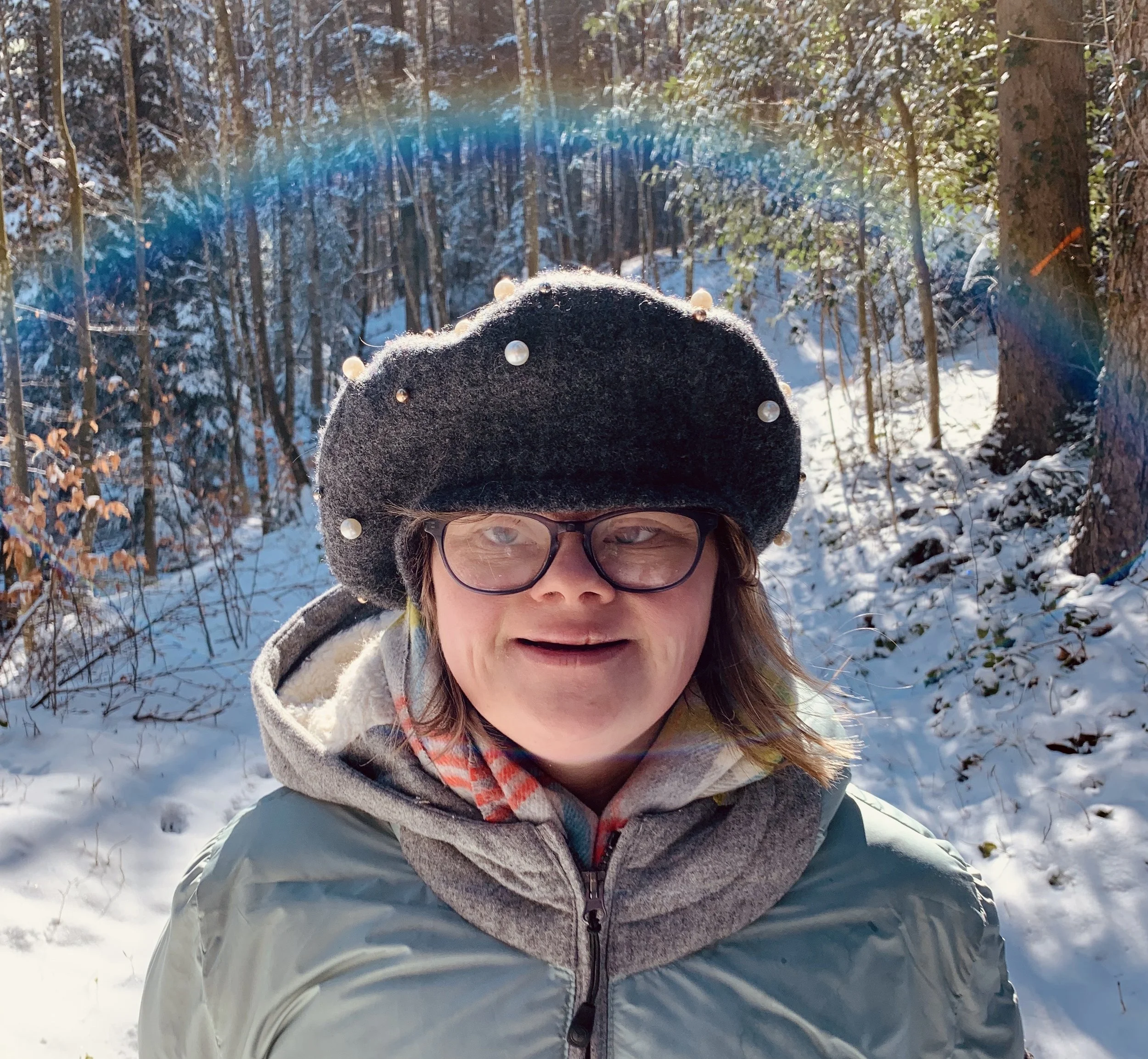 A woman wearing a black beanie decorated with pearl-like beads, glasses, and a gray jacket taking a selfie outdoors in a snow-covered forest with sunlight filtering through trees.
