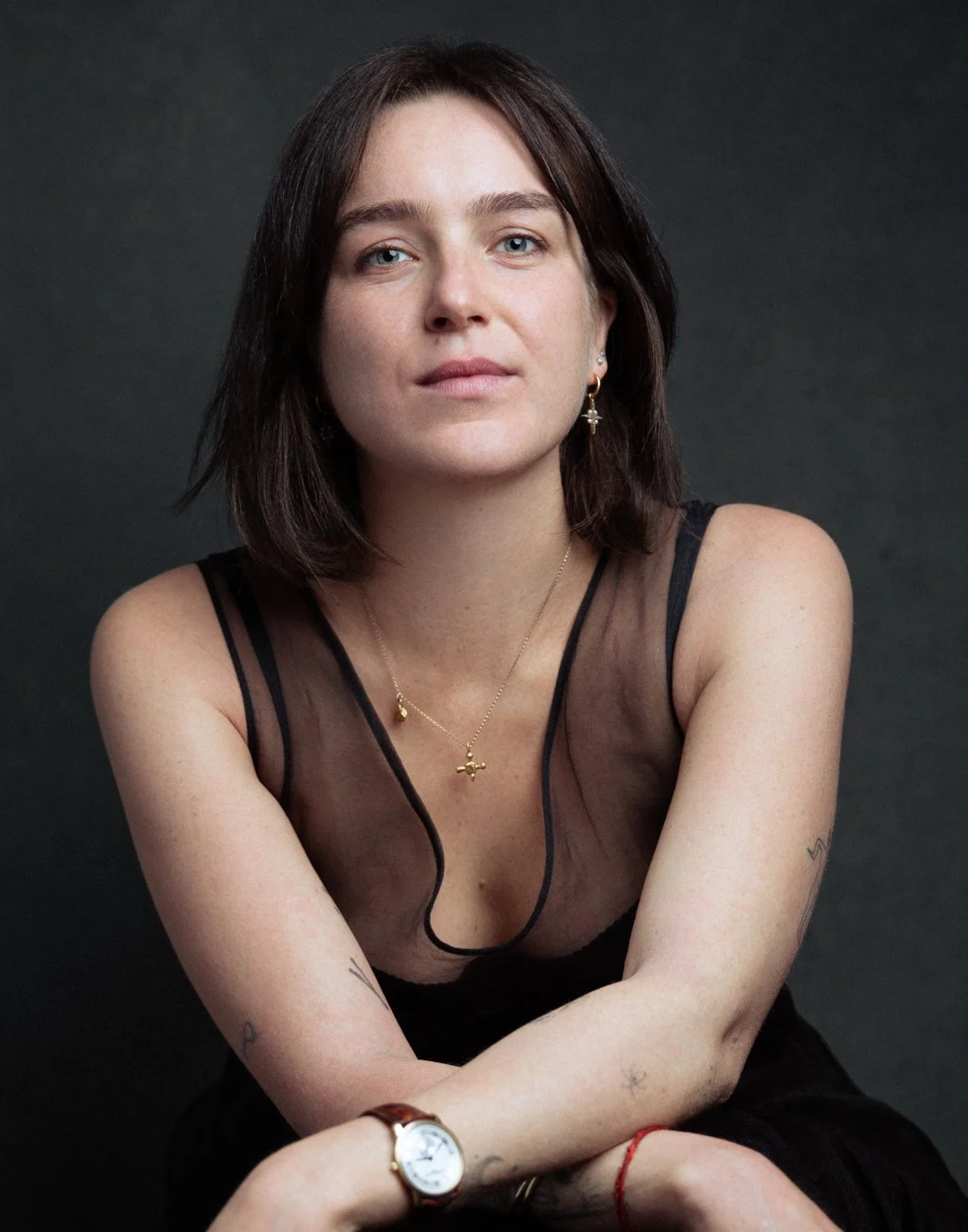 A woman with short dark hair and light skin, wearing a black sheer top, gold jewelry, and tattoos on her arms, posed against a dark background.