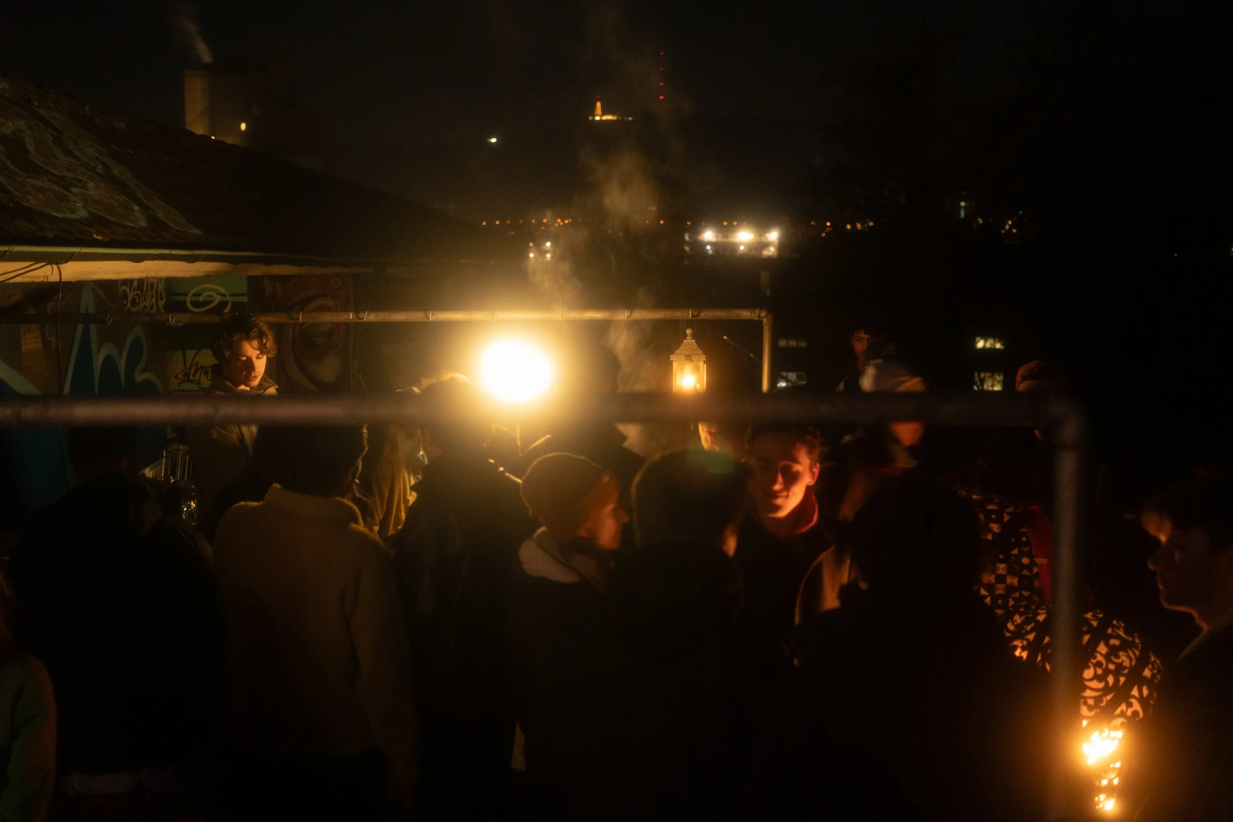 Night scene of a crowd gathered around a fire with people standing and chatting, city lights, and a smoky sky in the background.