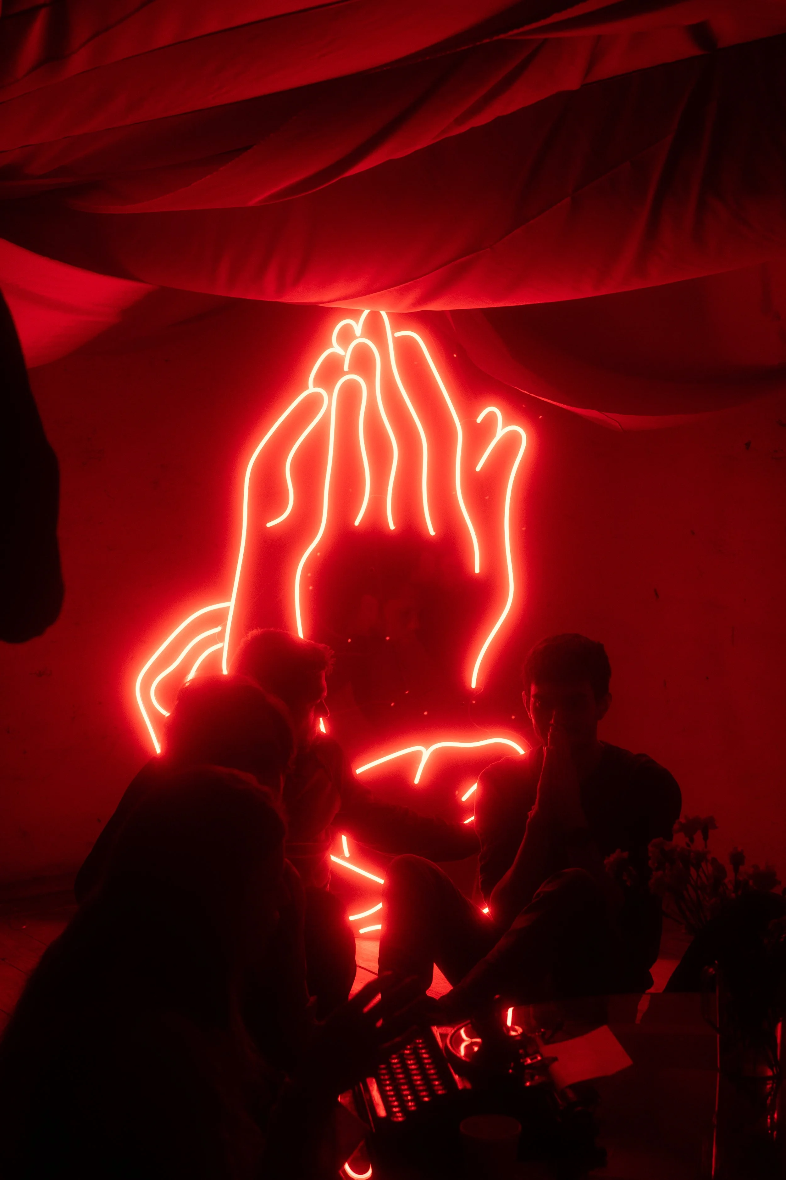 People sitting and standing in front of a red neon sign shaped like hands pressed together in a prayer or greeting gesture, with large red fabric drapery overhead.