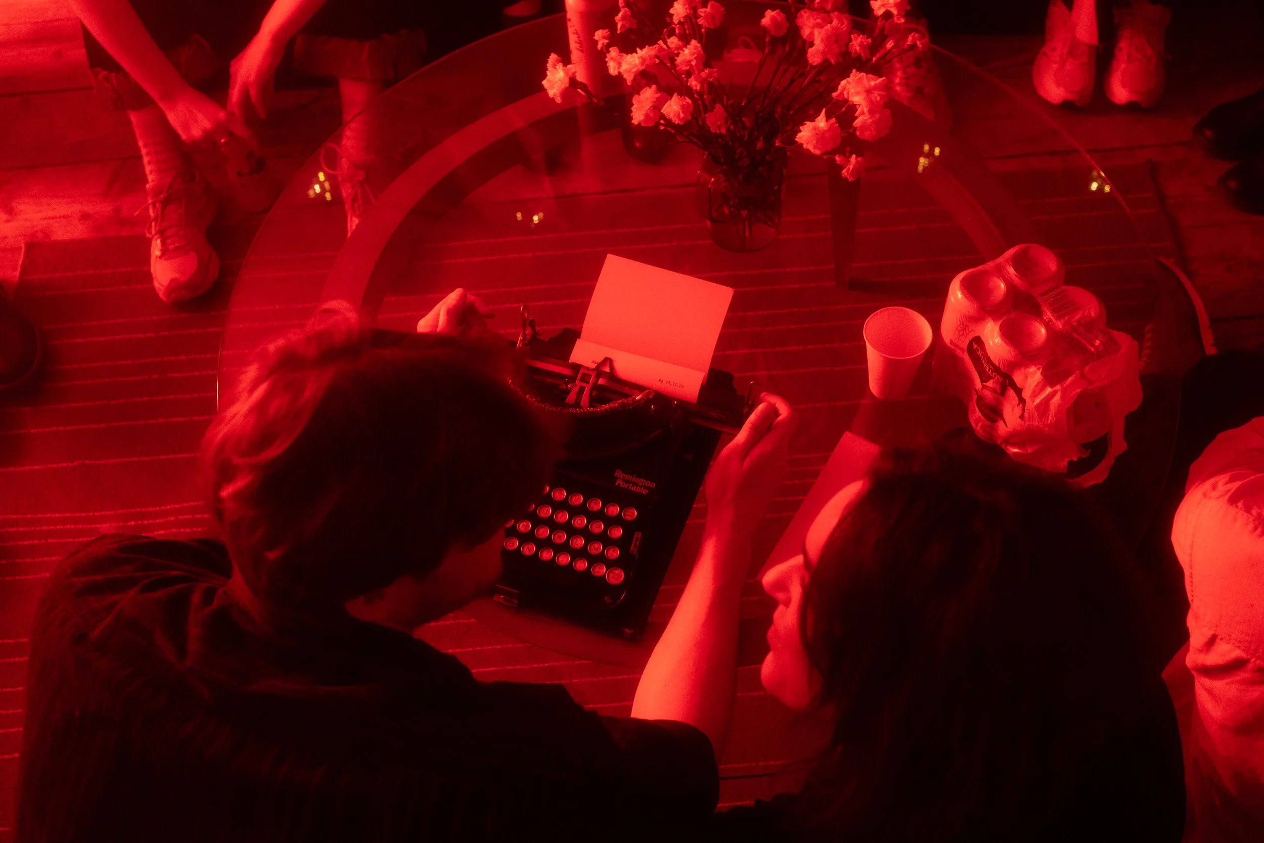 People sitting around a glass table with a vase of pink flowers, soda bottles, paper cups, and a small electronic device, illuminated by red lighting.