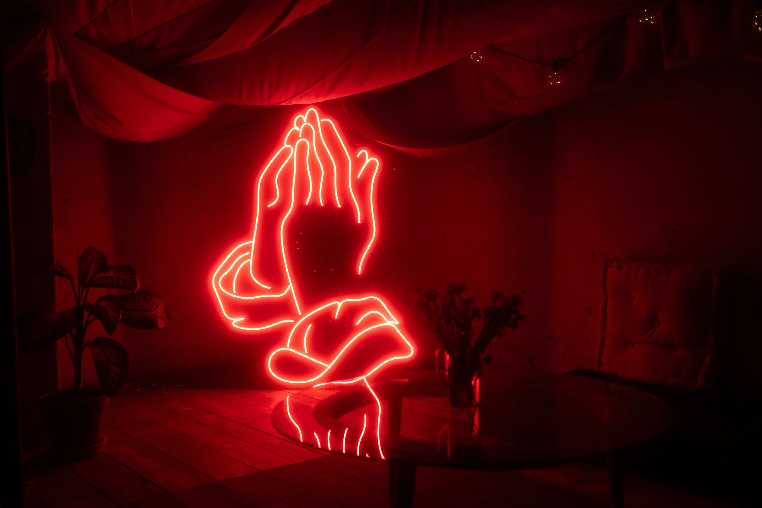 Neon sign of praying hands on a dark wall with red fabric draped from the ceiling and some plants nearby.