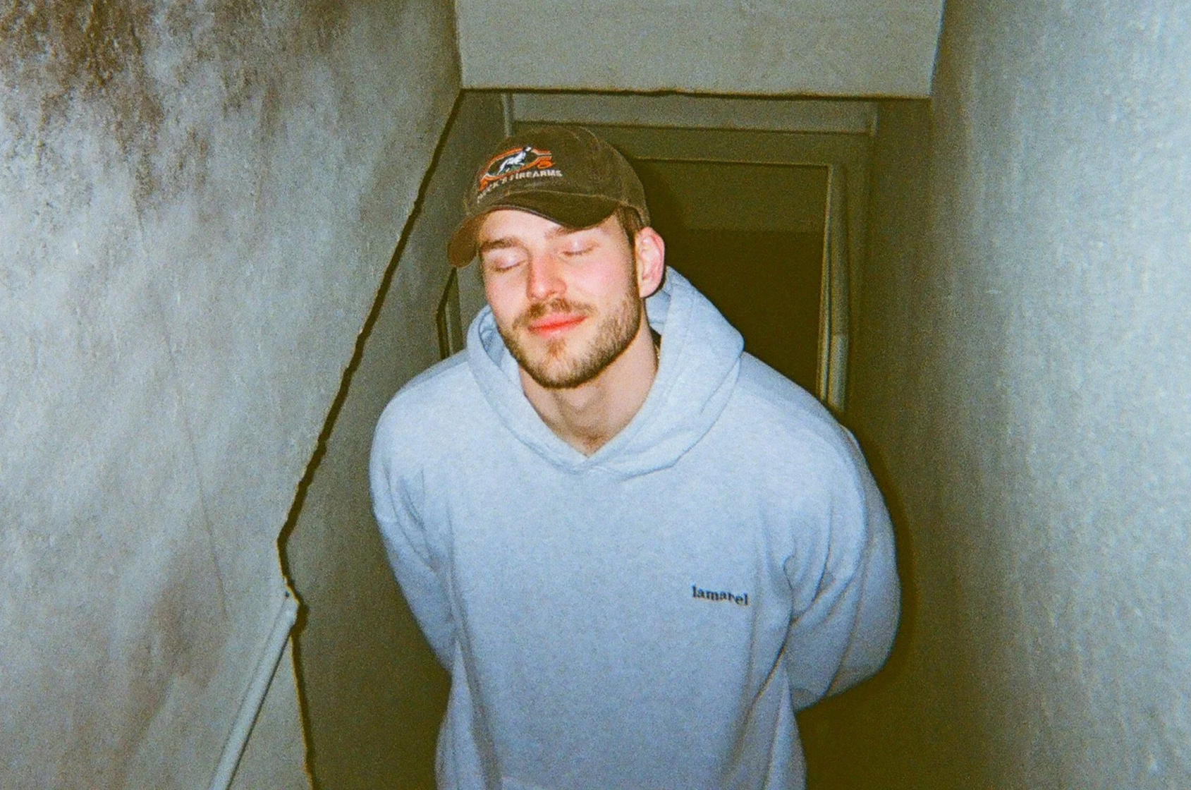 A young man with a beard and closed eyes, wearing a gray hoodie and a black cap, standing in a narrow, carpeted stairwell with beige textured walls.