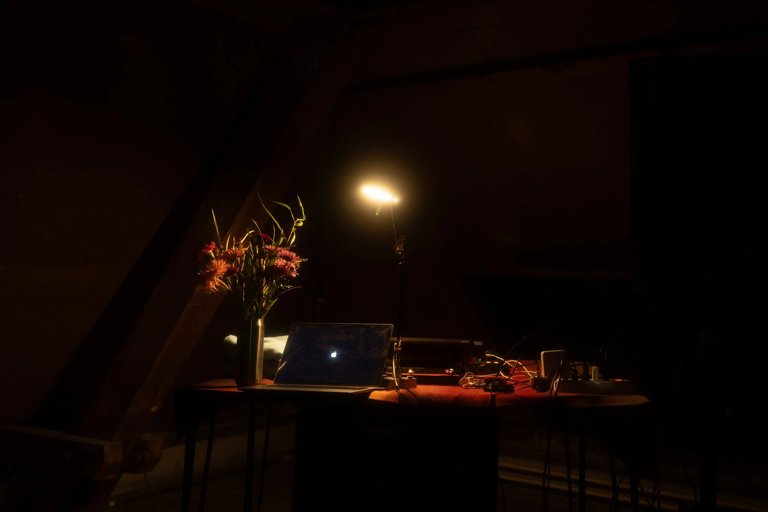 A dimly lit workspace with a laptop, a vase of colorful flowers, a lamp, and electronic equipment on a table.