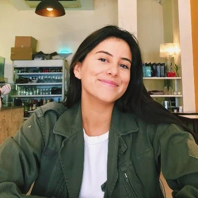 A woman with dark hair smiling at the camera, wearing a green jacket and white shirt, sitting in a café with shelves of drinks and a window in the background.
