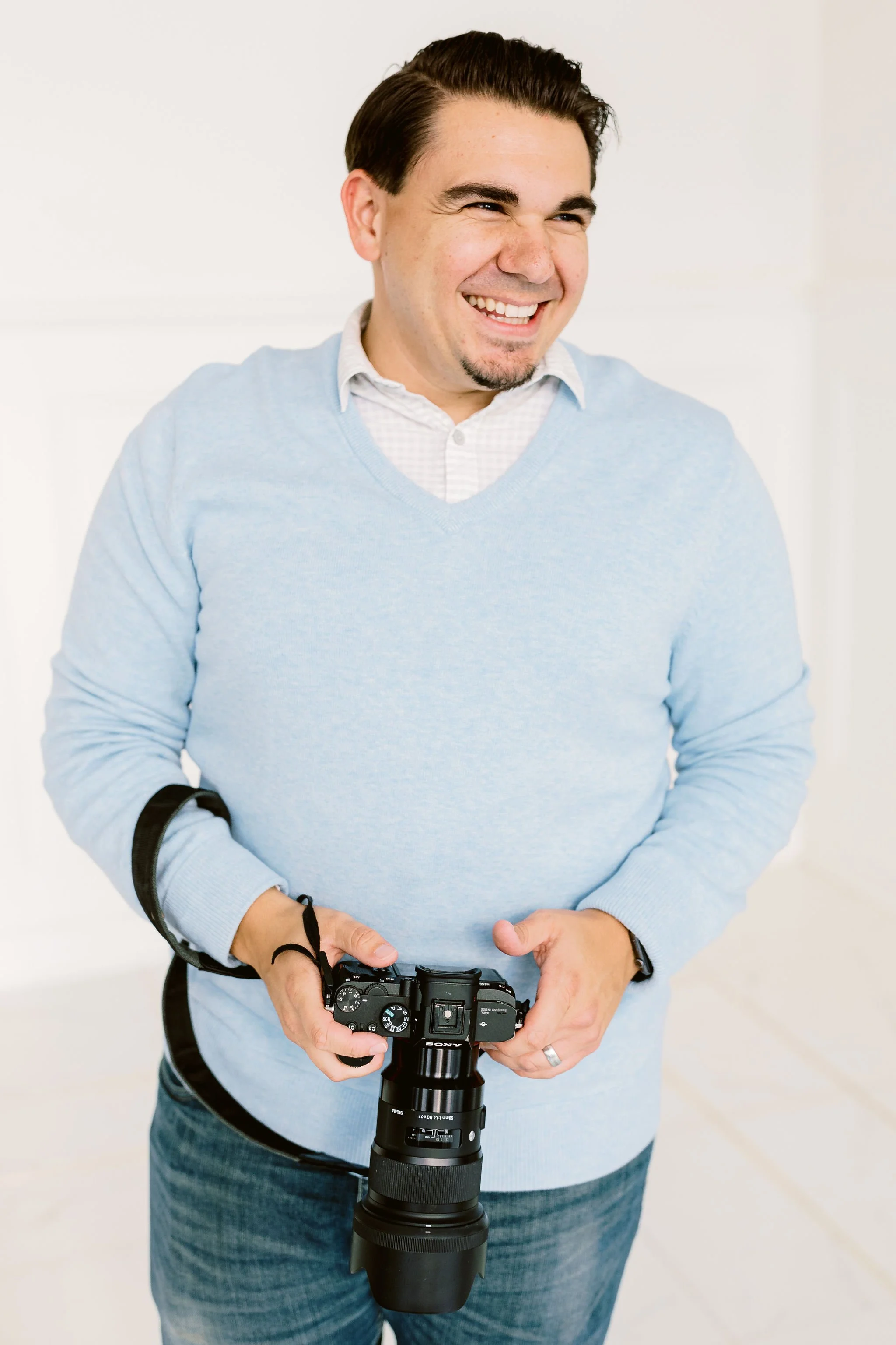 A man in a light blue sweater holding a camera, smiling, against a plain white background.