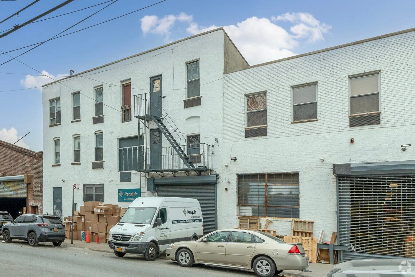 A white brick building with several windows, some with blinds, parked cars, a white delivery van with the Penguin logo, and cardboard boxes outside.
